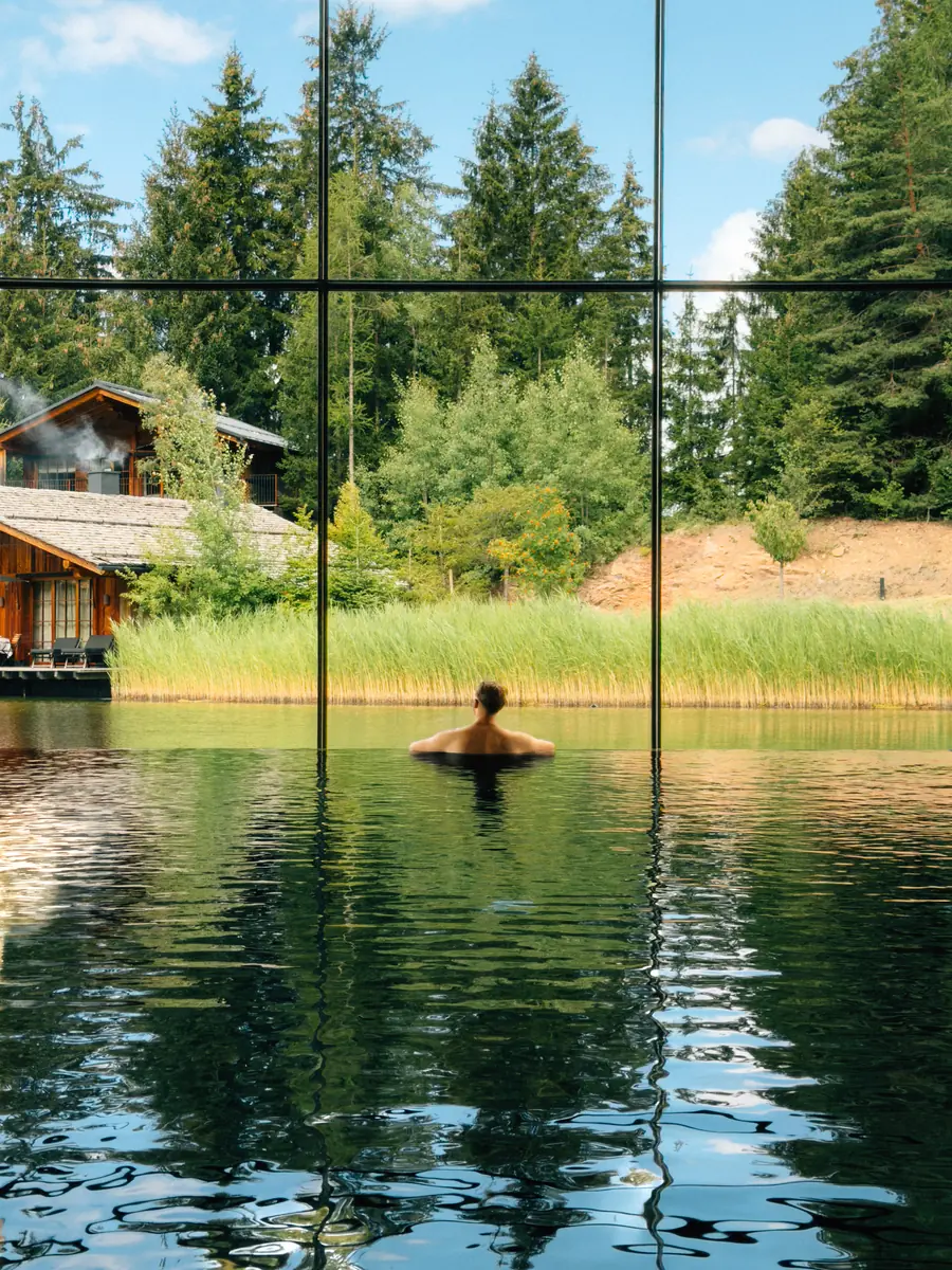A swimmer in an indoor pool facing a forest through tall windows